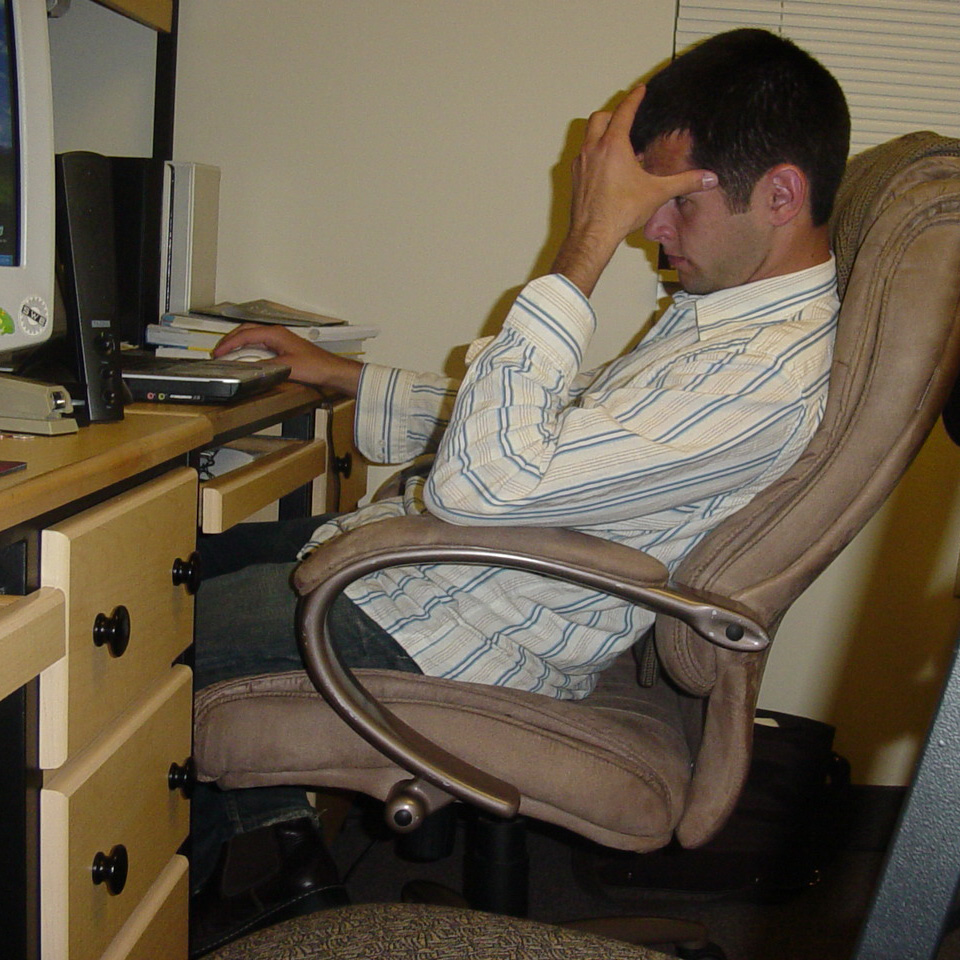 Hand on head, sitting in a chair, slouched, staring at a laptop.